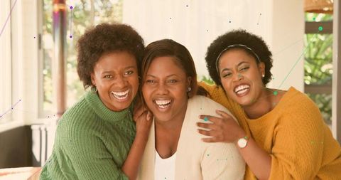 Laughing trio of mid-adult women hugging on couch in cozy living room with knit sweaters