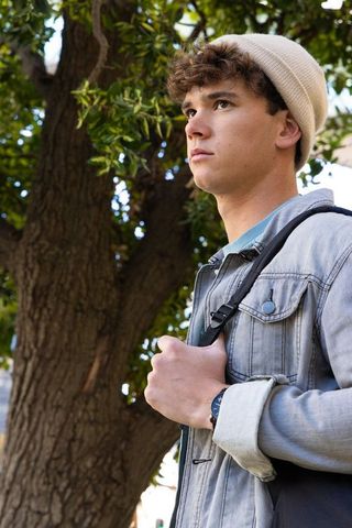 Thoughtful teen student wearing beanie and denim jacket holding backpack strap under tree