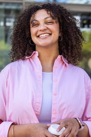Smiling african american woman holding coffee cup wearing pink shirt in modern urban plaza
