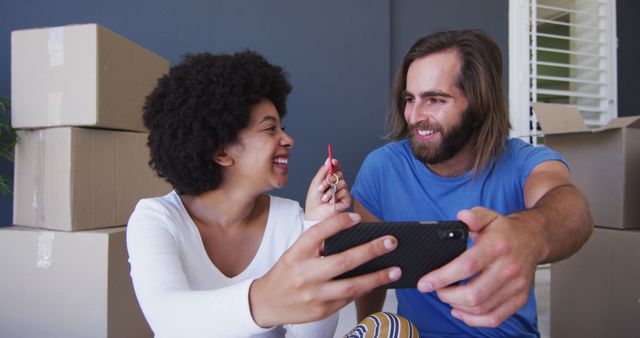 This image portrays a cheerful young couple celebrating their new home by taking a selfie surrounded by moving boxes. Perfect for content related to real estate, homeownership, moving day experiences, and lifestyle blogs or articles emphasizing the joy of starting a new chapter in life. It can also illustrate themes of accomplishment and personal milestones.