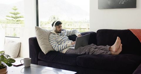 Indian man reclining on sofa using laptop and headphones at home in bright living room