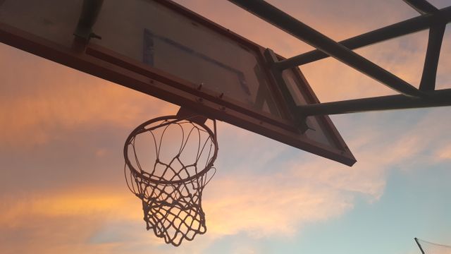 Basketball hoop silhouetted against a vibrant sunset sky, creating a serene sports atmosphere. Ideal for use in sports-themed designs, motivational posters, outdoor activity promotions, evening games advertising, and leisure sports campaigns.