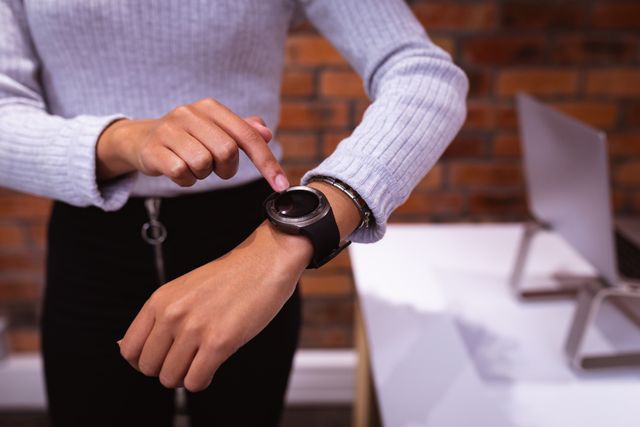 Front view mid section of a confident biracial businesswoman working in a modern creative office, wearing a lilac top and black skirt pointing at her smartwatch, brick wall in the background.