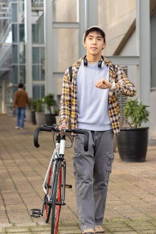 Young asian male walking road bike on campus checking smartwatch urban commuter lifestyle