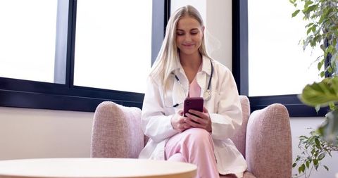 Smiling nurse sitting in sunlit lounge checking smartphone wearing white coat pink scrubs