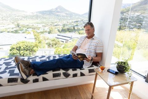 Mature man reclining on window seat reading paperback with mountain view and tea break