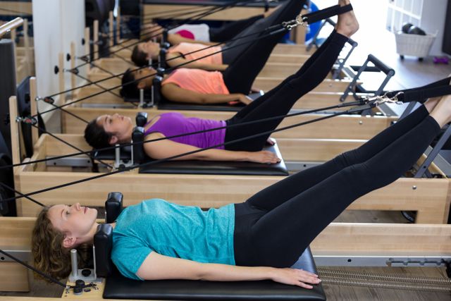 Group of women lying on reformer machines during a pilates class in a gym. The image can be used for promoting fitness centers, pilates classes, exercise equipment, and health-related articles. It conveys themes of fitness, group activities, and healthy lifestyle.