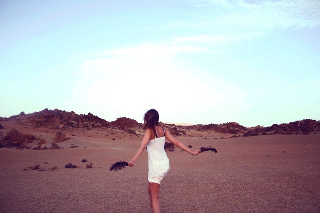 Woman in white dress holding black feathers in wide desert landscape. Ideal for themes of adventure, solitude, and summer exploration. Perfect for use in travel brochures, nature editorials, and inspirational blogs focused on freedom and nature's beauty.