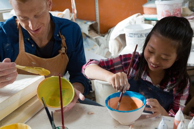 This image captures a young girl joyfully painting a bowl alongside an experienced potter in a vibrant, busy pottery workshop. Ideal for educational materials, art class promotions, DIY craft tutorials, and articles focusing on creative skills development and mentorship.