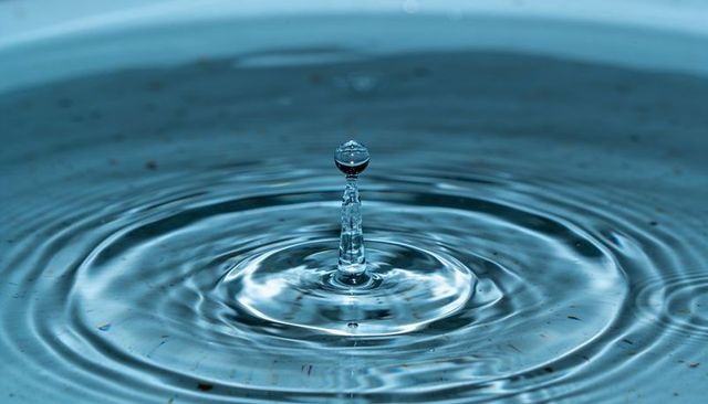 Crystal water droplet rising from rippled surface creating concentric rings and reflections