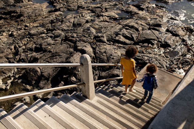 High angle rear view of a biracial woman wearing a yellow dress and her son enjoying time together by the sea, holding hands and walking down stairs from a promenade to the beach on a sunny day