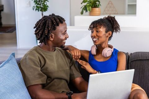 African american couple relaxing on sofa laughing with laptop and headphones in cozy home