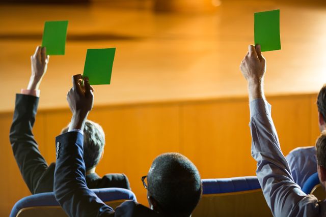 Business executives are raising green cards to show their approval during a conference. This image can be used to depict decision making, teamwork, and active participation in corporate events or seminars. Ideal for illustrating concepts related to leadership, collaboration, and professional gatherings.