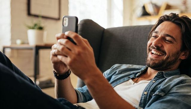 Smiling bearded man reclining on sofa holding smartphone in sunlit cozy living room