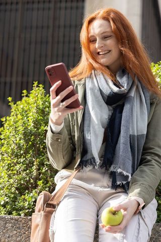 Redhead woman smiling while checking smartphone and holding apple on city ledge