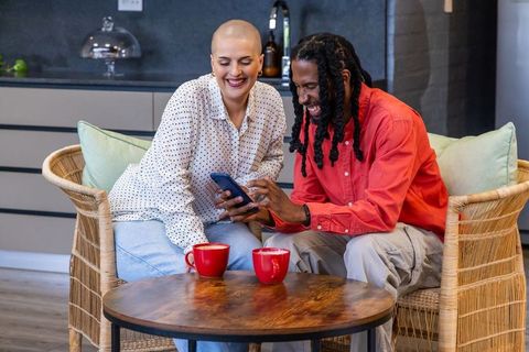 Couple sharing smartphone and red coffee mugs while laughing in cozy modern kitchen lounge