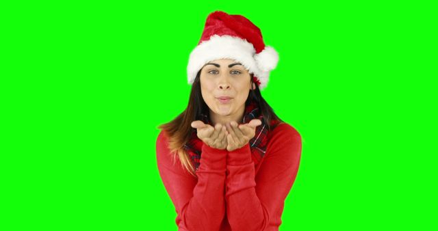 This vibrant image features a woman joyfully blowing a kiss while wearing a Santa hat and red sweater. Perfect for holiday-themed advertisements, festive social media posts, or any content celebrating the Christmas season. The green screen background provides versatility for custom backgrounds in various marketing materials.