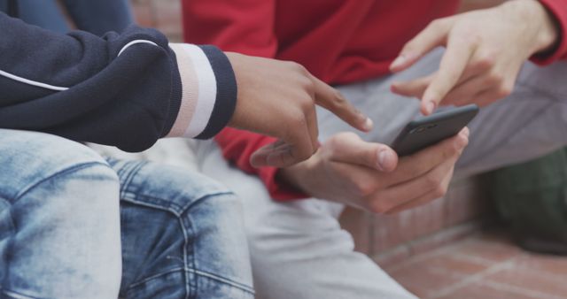 Two friends sitting outside using a smartphone, focusing on their hands and the device. Could be used to represent social interaction, technology use, mobile apps, or youth culture. Ideal for marketing campaigns, technology-related content, or educational materials highlighting digital communication.