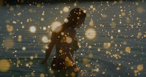 Wading woman at golden hour creating shimmering bokeh and ripples in calm sea