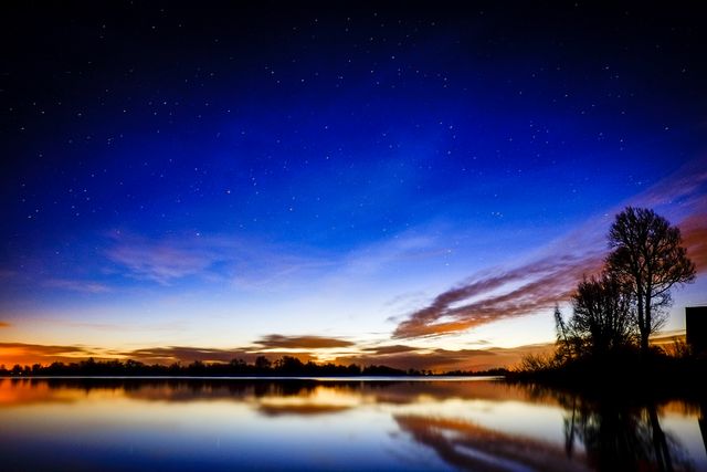 Photograph captures a breathtaking scene at twilight with a clear sky full of stars, reflected in a calm lake. Silhouettes of trees frame the horizon in the distance. Perfect for use in nature-themed publications, travel blogs, calming room decor, or inspiration for meditation and relaxation content.
