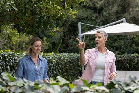 Senior woman guiding adult daughter through backyard garden pointing at plants and advising