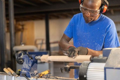 African american craftsman sanding wooden spindle on lathe wearing safety gear in workshop