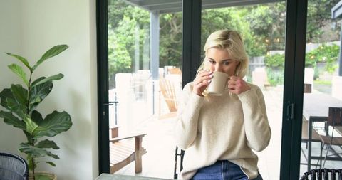 Blonde woman sipping coffee at sunlit table by patio doors, wearing cozy sweater