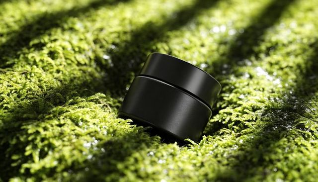 Matte black cosmetic jar resting on vibrant moss with dew and sunlit shadows minimal luxury mockup