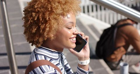Smiling young woman talking on smartphone while sitting on sunny urban steps