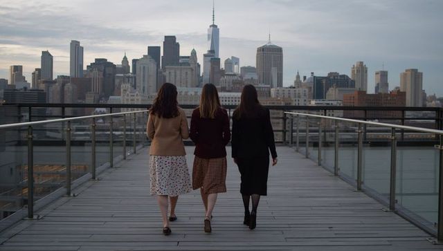 Three women walking toward manhattan skyline in smart casual coats on rooftop promenade