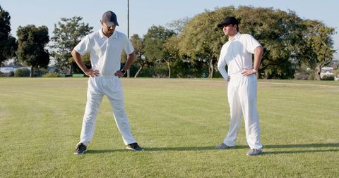 Two cricket teammates standing on sunlit grassy outfield wearing white uniforms and caps