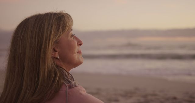 Middle-aged woman with blonde hair looking out into ocean during sunset. Scene evokes feelings of calm and reflection. Perfect for use in themes of meditation, mindfulness, relaxation, retirement, and happiness with nature.