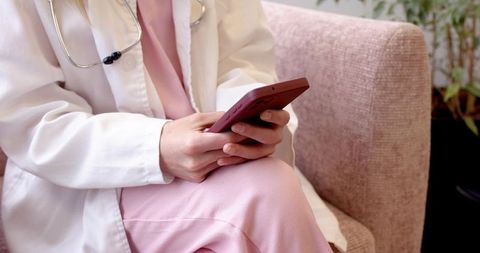 Clinician tapping smartphone in white coat and pink scrubs with stethoscope, seated
