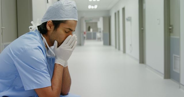 Healthcare professional in scrubs and surgical cap sitting in hospital hallway with head in hands, expressing exhaustion and burnout. Suitable for content on medical stress, healthcare worker fatigue, workplace well-being, hospital environment, and mental health awareness in the medical field.