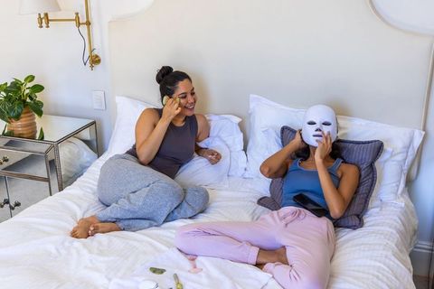 Multicultural friends relaxing in bed using jade roller and led face mask for skincare