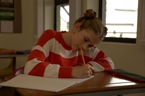 Focused schoolgirl writing in notebook at classroom desk with striped sweater and pencil