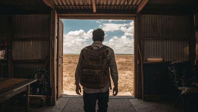 Standing backpacker framing vast arid plain from corrugated metal barn doorway, open sky