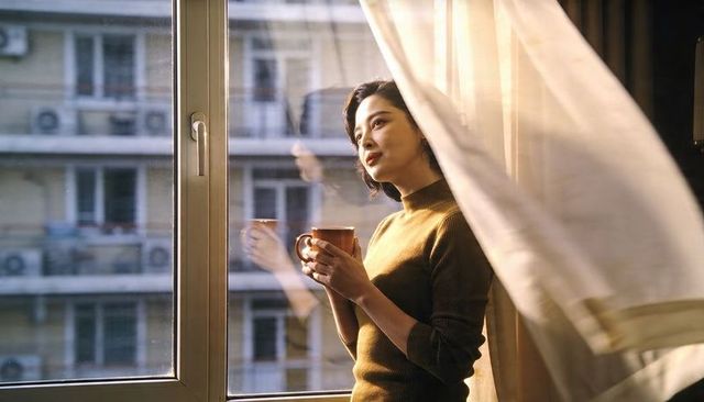 Asian woman standing by window holding mug, gazing out at city view with morning light