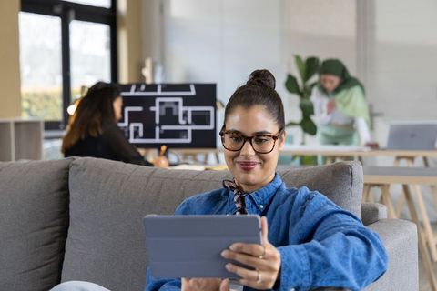Young professional woman using tablet on couch with collaborative open-plan team in studio