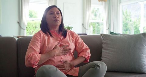 Asian woman holding chest and stomach while sitting on sofa at home, contemplative, sunlit