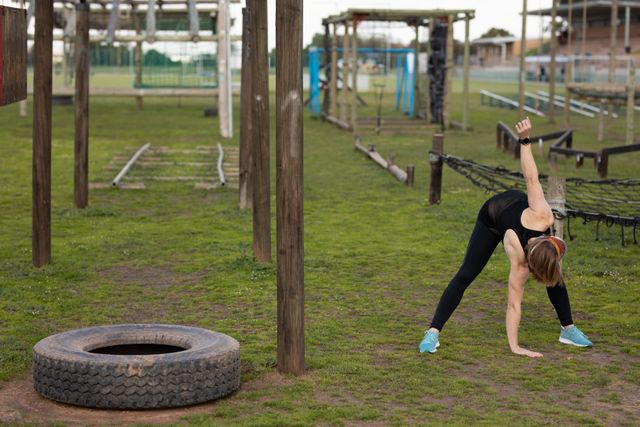 Caucasian woman in sports clothes stretching at an outdoor gym, preparing for a bootcamp training session. Ideal for use in fitness blogs, workout guides, health and wellness articles, and promotional materials for outdoor fitness programs.