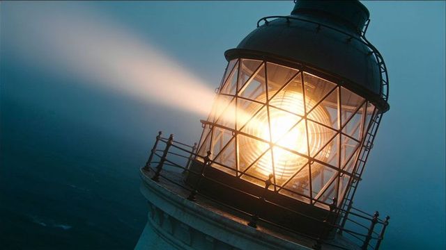 Historic lighthouse lantern room beaming fresnel lens through coastal fog with golden dusk light
