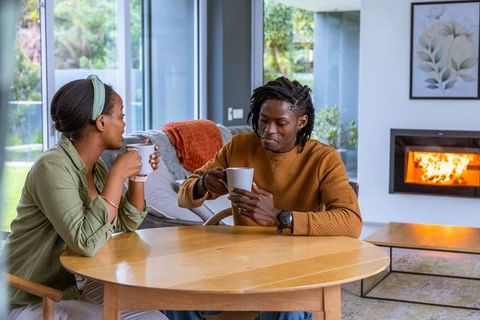 African american couple sipping coffee and chatting by fireplace in cozy living room