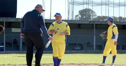Coach talking with baseball player holding bat on sunny diamond in yellow uniforms