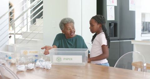 Grandmother teaching child recycling at kitchen table sorting paper and plastic bins