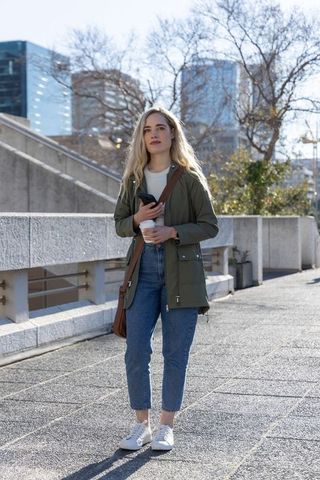 Young woman standing in city plaza with coffee and smartphone wearing olive parka