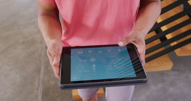 This image shows a man holding a digital tablet while controlling smart home features. The person is casually dressed in a pink shirt, engaged with a touchscreen device. Suitable for content related to smart technology, home automation, modern living, digital devices, and convenience. Great for blogs, articles, and advertisements focusing on technology integration in daily life.