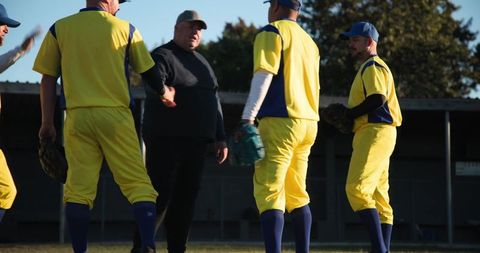 Veteran coach leading huddle with baseball team wearing yellow blue uniforms on field