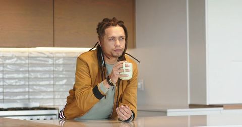 Thoughtful african american man leaning on modern kitchen island holding mug wearing mustard jacket