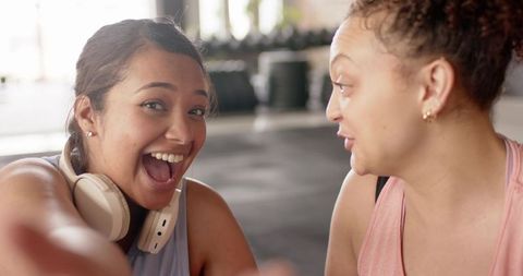 Two diverse women laughing and reaching forward in gym wearing headphones and workout tanks